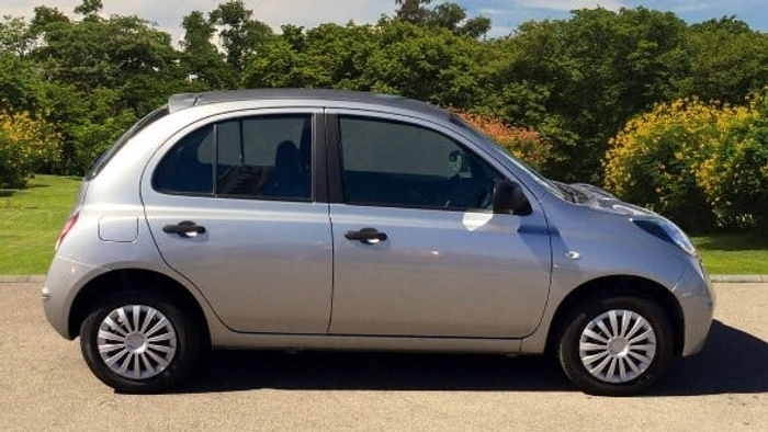 Silver compact used car parked on a paved surface with a background of green trees and bushes under a clear sky, suitable for new cars or used cars advertisements.