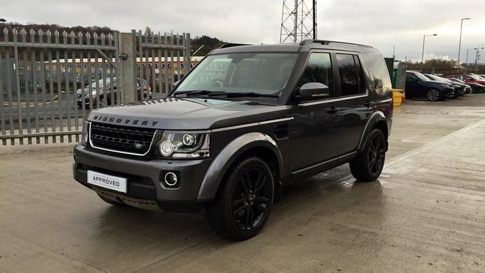 Grey Land Rover Discovery SUV parked on a concrete surface in a car lot with metal fencing and other vehicles in the background. The front grille displays the 'DISCOVERY' badge, and the number plate reads 'APPROVED'. The scene suggests a dealership environment featuring used cars and new cars for sale.