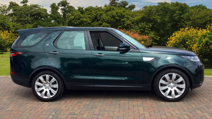 Green Land Rover Discovery SUV parked on a brick driveway with lush greenery and flowering bushes in the background, representing used cars and new cars in a suburban setting.