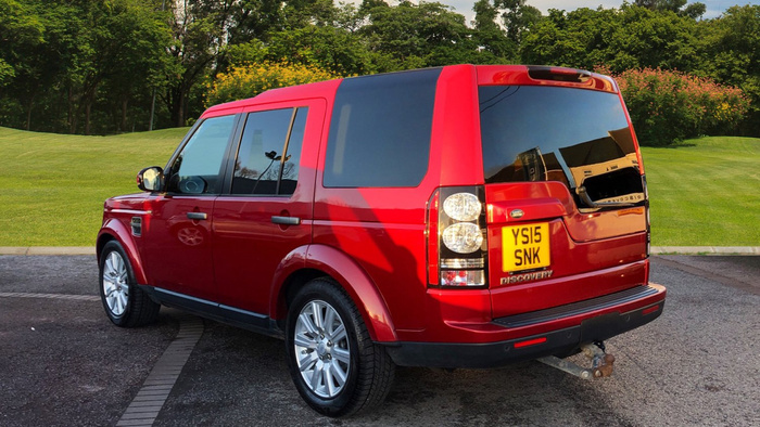 Red Land Rover Discovery parked on a paved surface with a green grassy area and trees in the background. The car has a yellow UK license plate reading YS15 SNK. The rear and right side of the vehicle are visible, showing tinted rear windows, silver alloy wheels, and a tow bar attached to the back. This image is suitable for used cars and new cars listings.