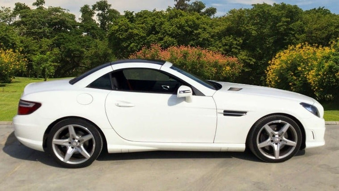 Side view of a white convertible sports car parked on a concrete surface with trees and bushes in the background, suitable for advertising used cars and new cars.