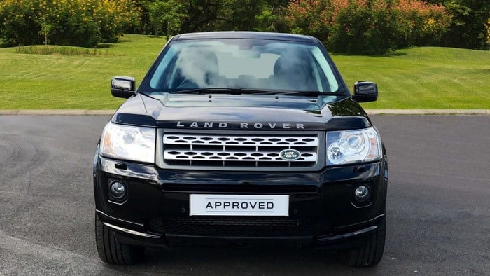 Front view of a black Land Rover parked on a driveway with green grass and bushes in the background. The car has the word 'LAND ROVER' displayed across the front hood and a licence plate with the word 'APPROVED'. This image relates to used cars and new cars.