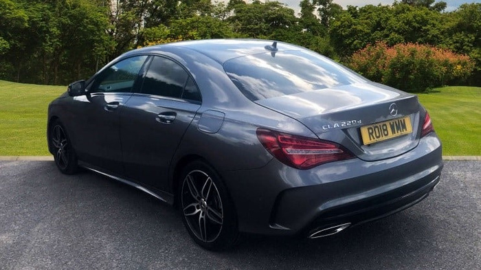 Rear side view of a grey Mercedes-Benz CLA 220d parked on a driveway with a green lawn and trees in the background. The car has a UK registration plate RO18 WMM, tinted windows, and sporty alloy wheels. This image features a stylish example of used cars, highlighting the sleek design and modern features of new cars in the Mercedes-Benz CLA series.
