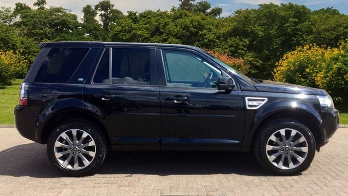Side view of a black Land Rover Freelander 2 parked on a paved driveway, surrounded by green bushes and trees in the background. The vehicle showcases alloy wheels and tinted windows, highlighting a well-maintained used car, suitable for buyers interested in used cars and new cars.