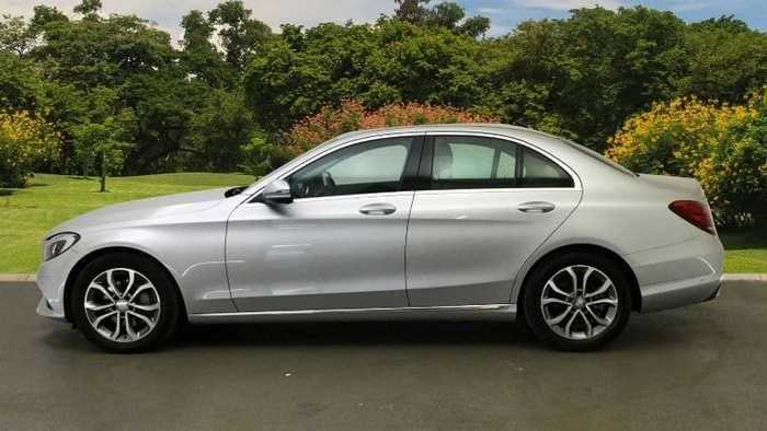Side view of a silver Mercedes-Benz sedan parked on a paved driveway with green trees and bushes in the background, representing a used car or new car available for sale.