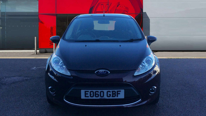Front view of a black Ford car parked on asphalt in front of a dealership with a red and grey exterior wall and the word 'Welcome' partially visible on the red panel. The car has a visible registration plate reading 'EO60 GBF'.