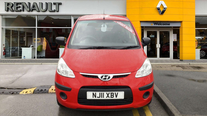Red Hyundai car with registration plate NJ11 XBV parked outside a Renault dealership with large Renault logo and signage. The car has a windscreen sticker reading 'Special Trade Sales'. The dealership entrance has a yellow facade and a welcome sign above the doors. Rain droplets are visible on the car's bonnet.