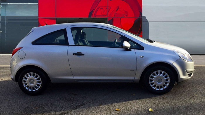 Side view of a silver three-door hatchback car parked in front of a building with a red and black logo featuring a griffin emblem. The car has steel wheels with hubcaps, a black door handle, and a fuel cap on the rear side. The background includes a modern building facade with a prominent red panel displaying the griffin logo, suggesting a car dealership specialising in used cars and new cars.