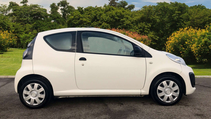 Side view of a small white three-door hatchback car parked on a tarmac surface with lush green trees and yellow flowers in the background, representing a used car or new car for sale.