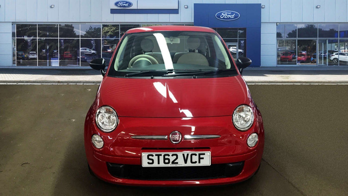 Red Fiat car with license plate ST62 VCF parked in front of a Ford dealership building with large glass windows and Ford logos. Reflection of cars visible inside the showroom through the windows. Scene highlights both used cars and new cars available at the dealership.