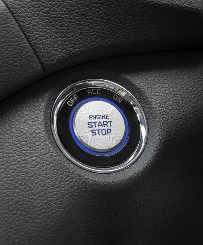 Close-up of a car's engine start stop button on a black textured dashboard. The button is circular with a metallic rim and has the text 'OFF', 'ACC', 'ON' around the edge, and 'ENGINE START STOP' in the centre, highlighting modern ignition technology used in new cars and available in used cars.