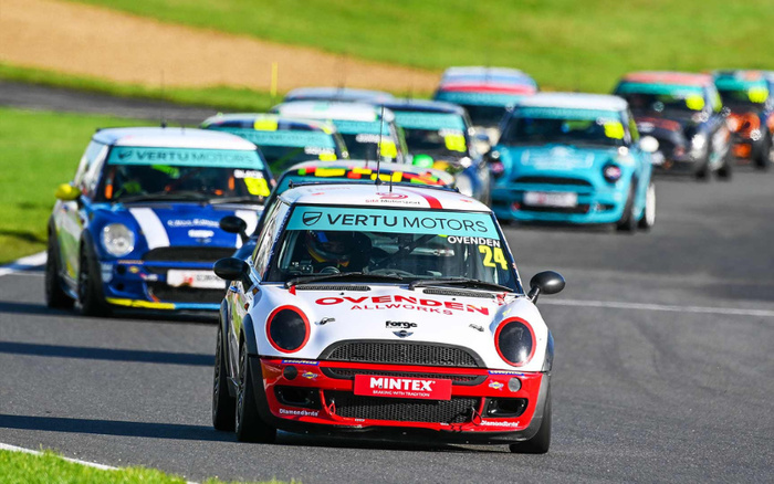 A group of race cars driving on a race track, featuring Mini Coopers with various sponsor logos. The leading white car with red accents displays 'VERTU MOTORS' and 'OVENDEN ALL WORKS' on the bonnet and windshield, along with the number 24. Other cars behind it also display 'VERTU MOTORS' branding. The image relates to used cars and new cars through the visible Vertu Motors sponsorship.
