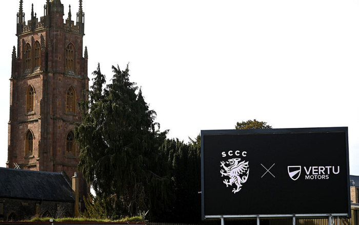 Black billboard with white logos and text showing SCCC with a lion emblem next to an 'X' and Vertu Motors logo, situated outdoors near a large historic stone church tower and tall trees under a clear sky, promoting the collaboration between SCCC and Vertu Motors, a dealership known for used cars and new cars.