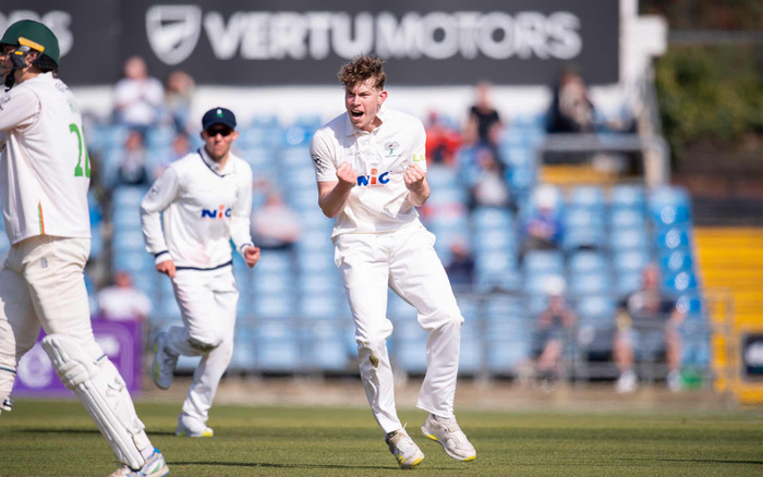 Cricket players on the field during a match, with one bowler celebrating a wicket. The players wear white uniforms with the logo 'NiC' on their chests. In the background, there is a blurred advertisement banner for 'VERTU MOTORS'. The image captures a moment of excitement in a cricket game, with emphasis on the branding related to Vertu Motors, a company associated with used cars and new cars.
