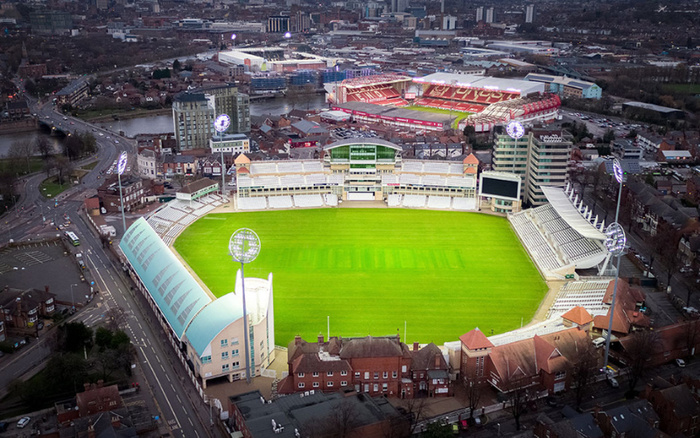Aerial view of a large empty cricket stadium with bright green pitch and seating stands, surrounded by city buildings and roads, featuring illuminated floodlights during dusk, with no visible cars or vehicles on site, emphasising the stadium structure and urban setting without direct connection to used cars or new cars.