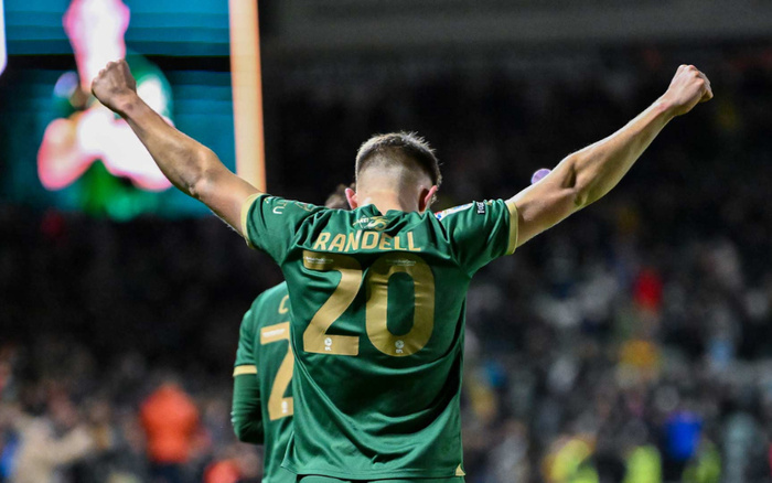 Football player wearing a green jersey with the name 'RANDELL' and number '20' celebrating with arms raised in a stadium filled with spectators at night, focusing on the player from behind.