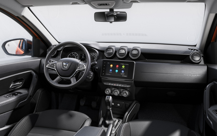 Interior view of a modern car focusing on the dashboard and steering wheel, featuring a digital display screen with infotainment options and air vents above it. The car has a black and grey colour scheme with leather seats and a gear shift lever in the centre console. The steering wheel displays a Dacia logo. This image is relevant for used cars and new cars showcasing interior design and technology.