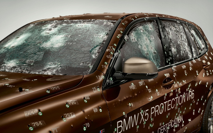 Close-up of a brown BMW X5 SUV with extensive simulated bullet damage and numbered markers on the body and windows, indicating impact points. Text on the car's side reads 'BMW X5 PROTECTION VR6' and 'CERTIFICATION'. The image suggests testing or demonstration of vehicle protection, relevant to used cars and new cars in terms of durability and safety features.