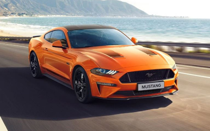 Bright orange Ford Mustang GT driving on a coastal road with mountains and sea in the background, showing the front and side of the new car model with a visible Mustang badge on the grille and license plate. Ideal image for used cars or new cars listings.
