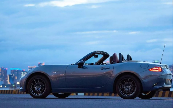 Side view of a modern dark grey convertible sports car parked on a road with city skyline in the background during dusk, highlighting sleek design and alloy wheels, ideal for showcasing used cars and new cars for sale.