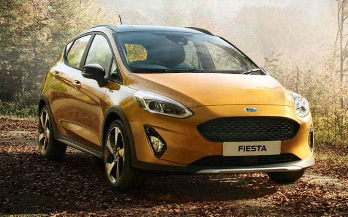 Golden yellow Ford Fiesta parked on a forest path with autumn leaves on the ground and trees in the background. The car is shown from the front-left angle with clear headlights, black grille, and a white license plate displaying the word 'FIESTA'. Ideal image for used cars and new cars listings.