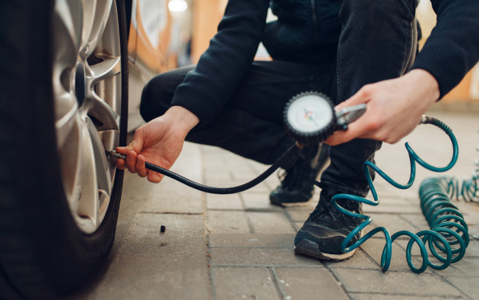Close-up of a person checking and inflating a car tyre using a manual tyre pressure gauge and an air hose on a paved surface, relevant for maintenance of used cars and new cars.