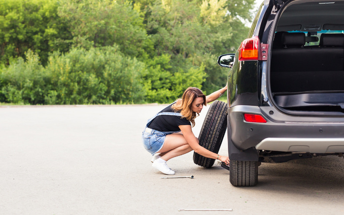 A woman changing the tyre of a black SUV parked on a paved area with green trees in the background. She is squatting and placing a spare tyre while using a wrench to remove the old tyre from the vehicle. The rear door of the SUV is open, displaying the empty boot space. This image relates to used cars and new cars with a focus on vehicle maintenance.