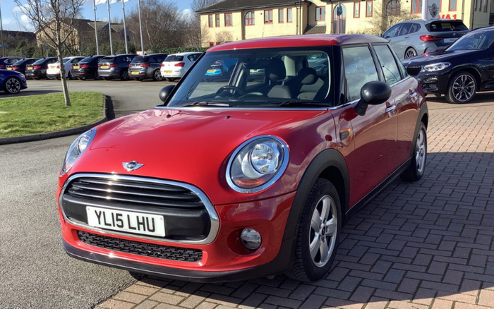 Red Mini Cooper car with registration plate YL15 LHU parked on a paved area in front of a car dealership lot, surrounded by other vehicles including a black BMW and a grey car, under a clear blue sky, showcasing options for used cars and new cars.