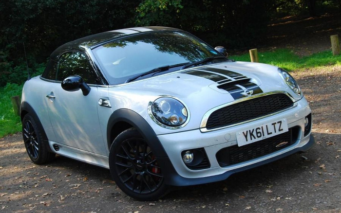 Silver Mini Cooper convertible car with black racing stripes and black alloy wheels, parked on a leaf-strewn path with greenery and trees in the background, displaying UK registration plate YK61 LTZ, suitable for used cars and new cars listings.