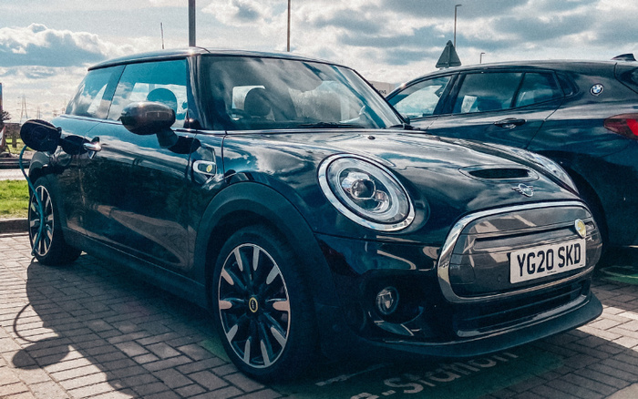 Black Mini Cooper with licence plate YG20 SKD parked on a paved lot next to a blue BMW. The scene shows a sunny day with partly cloudy skies, highlighting the exterior detail of these used cars, suitable for customers interested in both used cars and new cars.