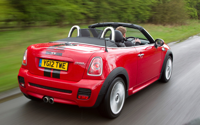 Red Mini Cooper S convertible driving on a wet road with a visible UK license plate YG12 TWE, featuring black racing stripes on the rear. The driver is a man with short blond hair. The car is surrounded by green trees and bushes, showcasing a sporty and compact used car, suitable for customers interested in both used cars and new cars.