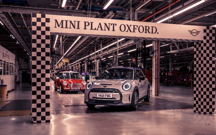 MINI Plant Oxford car manufacturing facility showing a white new MINI car with license plate YH21 FWG and a red used MINI car with license plate Y304 DBL under a checkered archway inside the factory. The image highlights the production and quality control of new cars and used cars within the automotive plant.