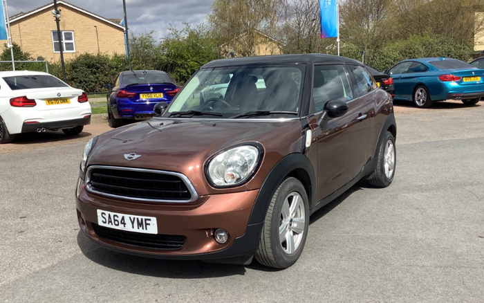 Brown Mini Cooper parked in a car lot with registration plate SA64 YMF, surrounded by various other vehicles including white, blue, and black cars; scene taken outdoors on a cloudy day with buildings and trees in the background, suitable for used cars and new cars market.