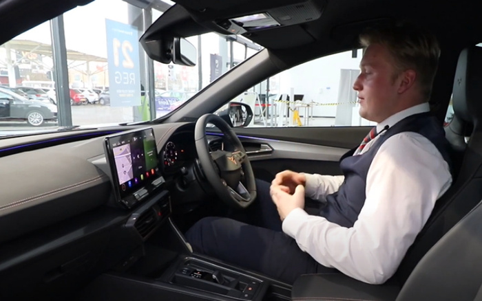 Interior view of a modern car featuring a digital touchscreen display and steering wheel with CUPRA logo, with a man in formal attire sitting in the driver's seat inside a dealership. Outside the window, a dealership sign reads '21 Reg' referring to used cars, and several cars are visible, indicating a showroom for both used cars and new cars.