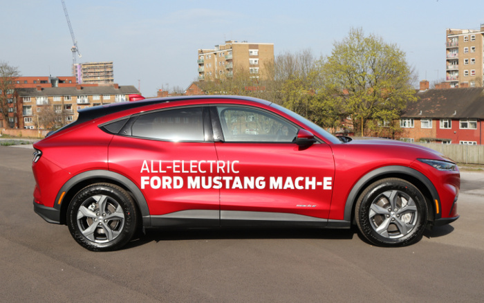 Side view of a red all-electric Ford Mustang Mach-E parked on a street in an urban area with residential buildings in the background. The car features bold white text on the door reading 'ALL-ELECTRIC FORD MUSTANG MACH-E'. This image highlights a new car model, suitable for advertisements related to used cars and new cars.