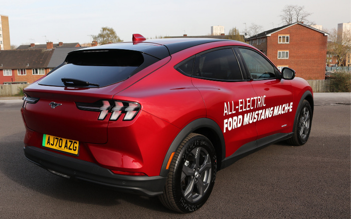 Red all-electric Ford Mustang Mach-E parked in a residential area, displaying text on the side panel reading 'ALL-ELECTRIC FORD MUSTANG MACH-E'. The car features a sleek design with tinted rear windows, modern rear lights, and a UK license plate AJ70 AZG, symbolising new cars available alongside used cars.