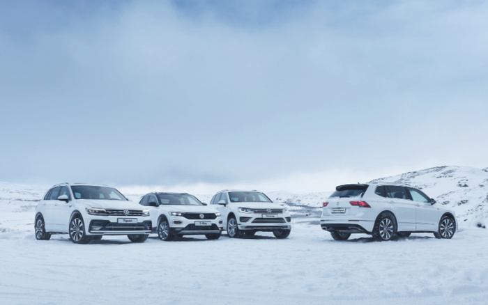 Four white Volkswagen cars parked in a snowy landscape with mountains in the background. The visible model names are Tiguan and T-Roc. The cars are clean and new looking, representing a range of new cars and used cars suitable for winter conditions.