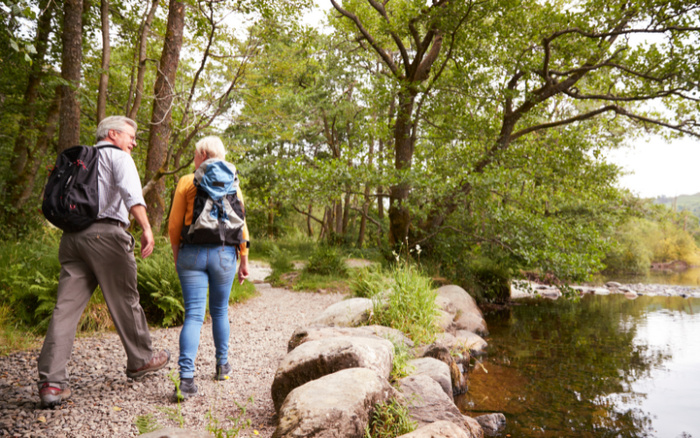 An older couple hiking on a gravel path along a serene river surrounded by lush green trees and rocks, both carrying backpacks, enjoying nature on a sunny day.