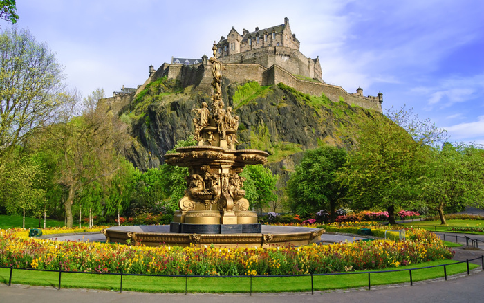 Ornate fountain surrounded by colorful flowers and green trees in a park with Edinburgh Castle on a rocky hill in the background under a clear blue sky, no visible used cars or new cars in the image.