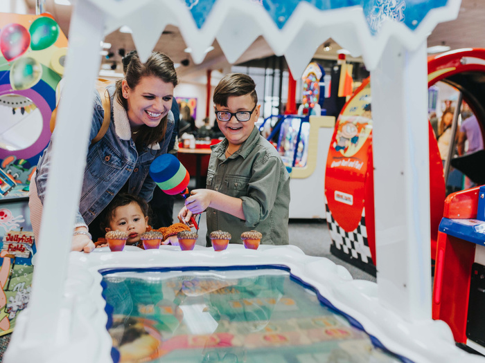 A smiling woman and two children playing an arcade game with colourful mallets in a bright indoor amusement centre, surrounded by various arcade machines and vibrant decorations, emphasising family fun and entertainment, not related to used cars or new cars.