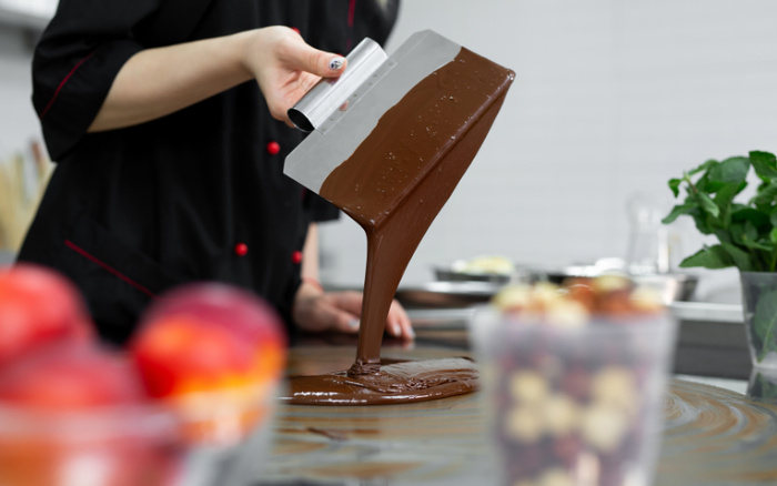 Person in a black chef coat spreading melted chocolate on a flat surface using a metal spatula, with blurred fruit and a glass container of chocolate-covered nuts in the foreground, and a green plant in the background in a modern kitchen setting, unrelated to used cars or new cars.