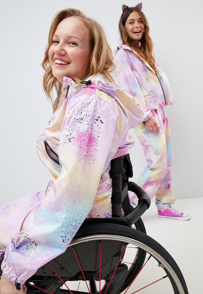 Two smiling women wearing matching pastel tie-dye tracksuits with pink, purple, blue, and yellow splashes; one woman with blonde hair sits in a wheelchair, and the other woman with long brown hair and cat ear headband stands behind her wearing pink sneakers, set against a plain white background. No text present. Optimised for used cars, new cars.