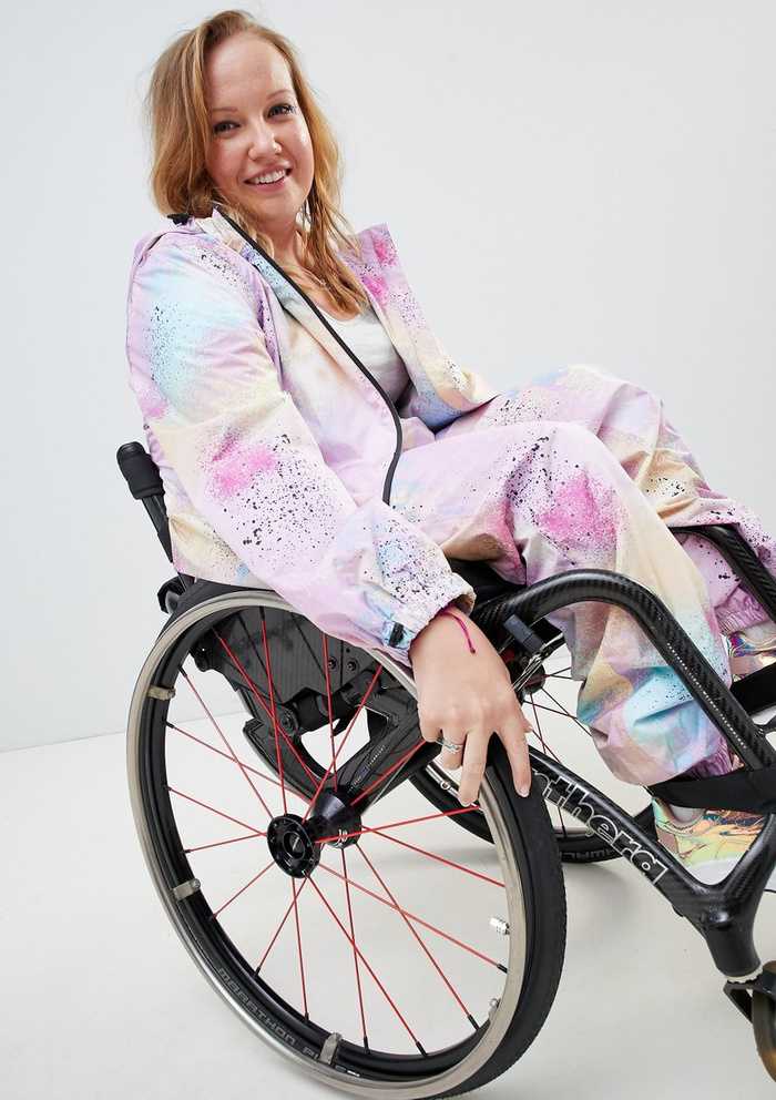 Young woman with light brown hair wearing a pastel-coloured splatter paint tracksuit sitting in a wheelchair with black frame and red spokes, smiling at the camera against a plain light grey background