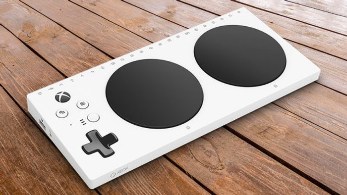 White Xbox Adaptive Controller with two large black circular buttons and various smaller buttons including a directional pad, placed on a wooden surface. The controller features the Xbox logo and the text 'XBOX' printed on the side. Image focused on the device without references to used cars or new cars.