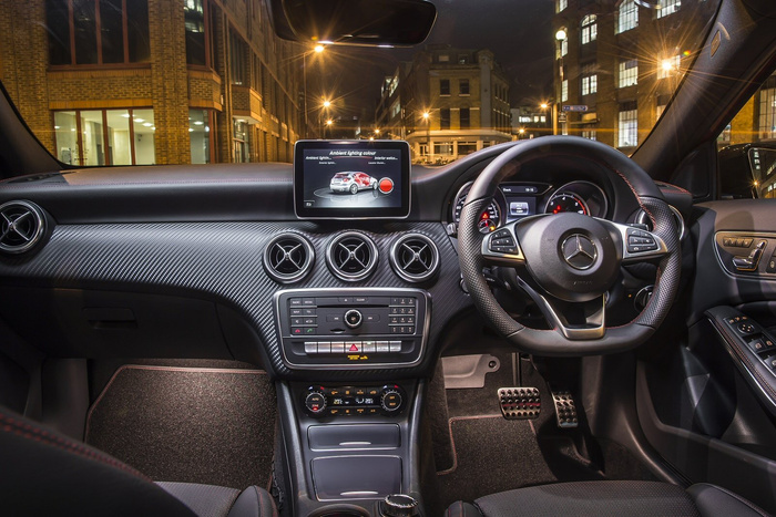 Interior view of a Mercedes-Benz car at night showing the dashboard with carbon fibre finish, three circular air vents, and a central multimedia display screen set to ambient lighting colour settings for exterior and interior lights. The steering wheel features the Mercedes logo, control buttons, and a digital instrument cluster is visible behind it. The centre console includes buttons for radio, media, and navigation, along with climate control dials displaying 20°C settings. The scene represents a modern, high-quality car interior typical of new cars available for purchase, relevant for used cars and new cars buyers.