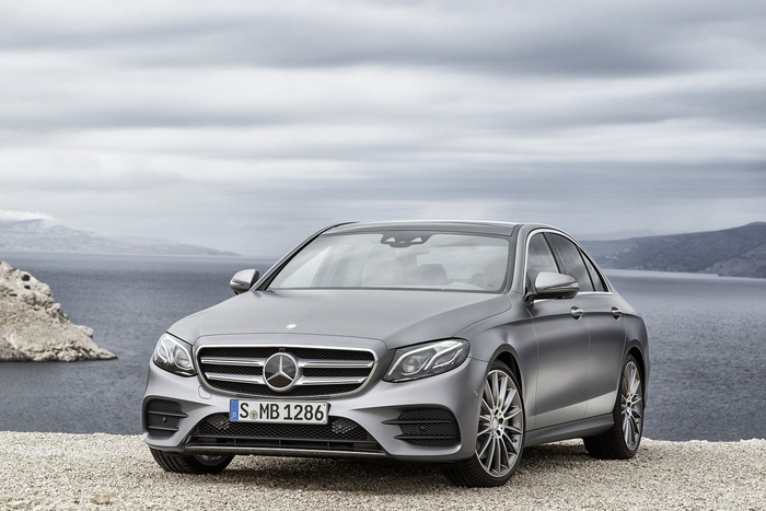 Matte grey Mercedes-Benz sedan parked on a gravel surface with a scenic coastal background under a cloudy sky, featuring the front grille with Mercedes emblem and German license plate reading 'S MB 1286', suitable for advertising used cars and new cars.