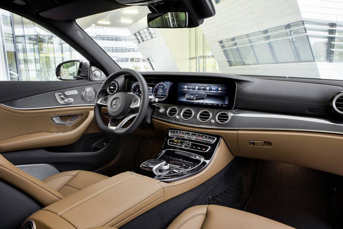 Interior of a luxury car featuring tan leather seats and door panels, a black and carbon-fibre dashboard, a multifunctional steering wheel with the Mercedes-Benz logo, and a digital instrument cluster and central infotainment screen displaying vehicle settings. The car is parked indoors with large glass windows showing a modern building exterior. This high-end interior exemplifies the quality found in new cars and is relevant for used cars buyers seeking premium features.