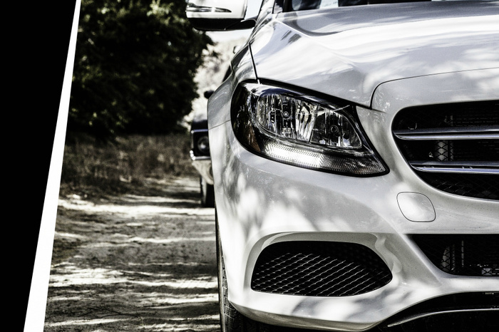 Close-up of the front left side of a white modern car, showing the headlight, grille, and side mirror with a blurred green and brown natural background on a dirt road, representing used cars and new cars.