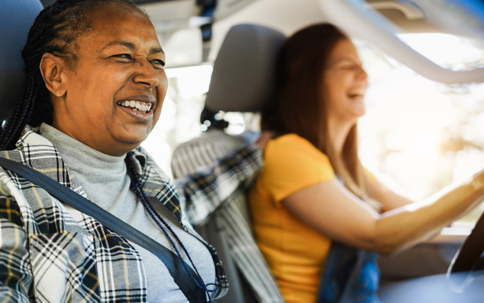 Two women happily sitting in a car, the driver in a yellow top and the passenger in a plaid jacket and grey turtleneck, both wearing seatbelts, enjoying a drive together, representing the joy of choosing used cars or new cars for a memorable travel experience.