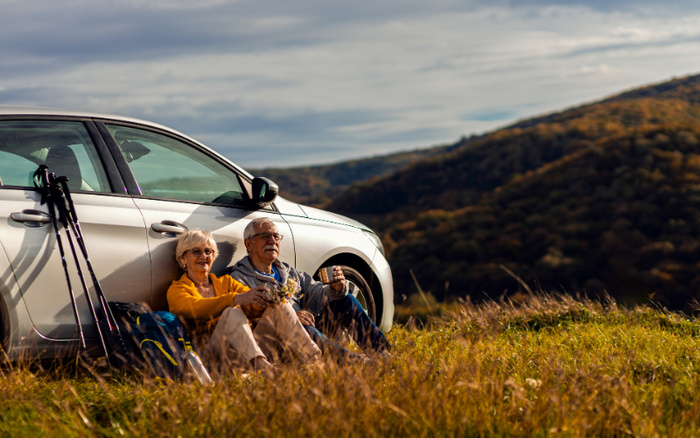 Senior couple sitting on grass leaning against a silver car in a scenic hill landscape, enjoying drinks and relaxation during a countryside outing, illustrating the comfort and freedom provided by used cars and new cars for outdoor adventures.